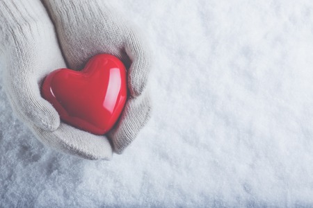 Female hands in white knitted mittens with a glossy red heart on a snow winter background.の写真素材