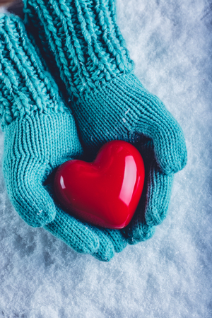 Woman hands in light teal knitted mittens are holding a beautiful glossy red heart in a snow winter background.の写真素材