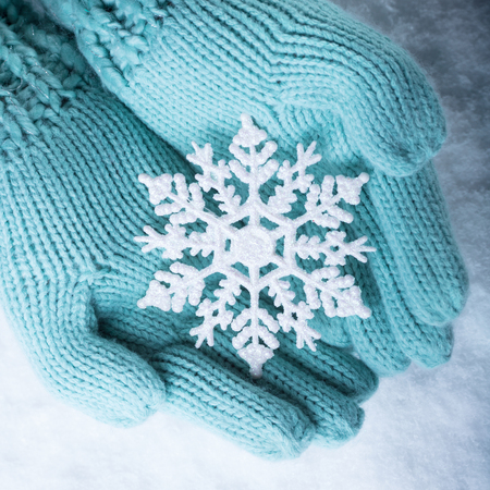 Female hands in light teal knitted mittens with sparkling wonderful snowflake on a white snow background.の写真素材