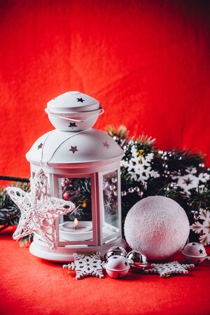Magical white lantern is standing with beautiful white knit star on it and a fir tree branch and a snowball lying in front of it on a christmas red background.の写真素材
