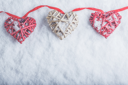 Three beautiful romantic vintage hearts are hanging on a red band on a white snow winter background.の写真素材