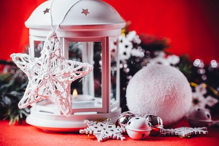 Magical white lantern is standing with beautiful white knit star on it and a fir tree branch and a snowball lying in front of it on a christmas red background.の写真素材