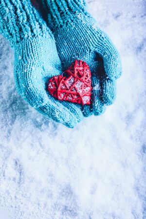 Woman hands in light teal knitted mittens are holding a beautiful entwined vintage romantic red heart in a snow background.の写真素材