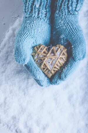 Woman hands in light teal knitted mittens are holding a beautiful entwined vintage romantic red heart in a snow background.の写真素材