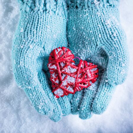 Woman hands in light teal knitted mittens are holding a beautiful entwined vintage romantic red heart in a snow background.の写真素材