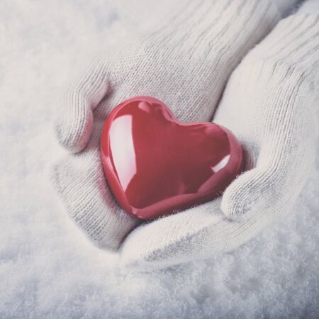 Female hands in white knitted mittens with a glossy red heart on a snow background.の写真素材