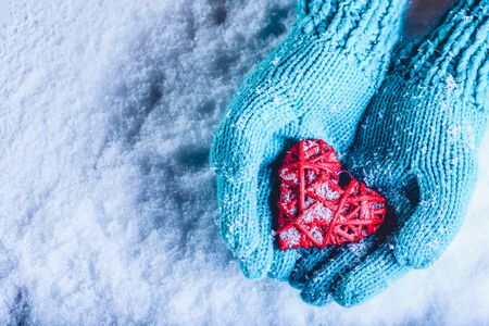 Woman hands in light teal knitted mittens are holding a beautiful entwined vintage romantic red heart in a snow background.の写真素材