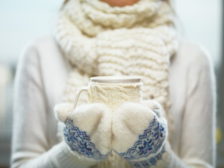  Woman hands in white and blue mittens holding a cozy knitted cup with hot cocoa, tea or coffee.