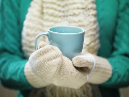 Woman hands in white woolen mittens holding a cozy cup with hot cocoa, tea or coffee.