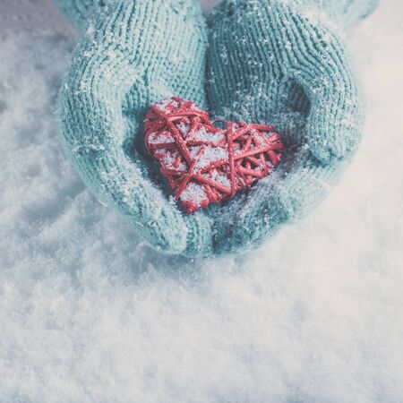 Woman hands in light teal knitted mittens are holding a beautiful entwined vintage romantic red heart in a snow background.の写真素材