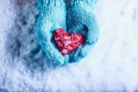 Woman hands in light teal knitted mittens are holding a beautiful a entwined vintage romantic red heart in a snow background.の写真素材