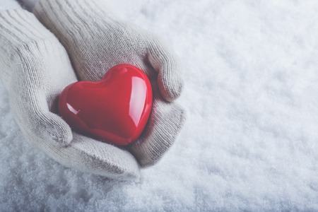 Female hands in white knitted mittens with a glossy red heart on a snow winter background.の写真素材