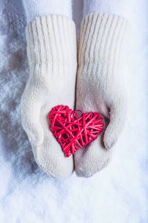 Female hands in white knitted mittens with a entwined vintage romantic red heart on a winter snow backgroundの写真素材