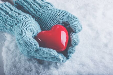 Female hands in light teal knitted mittens with a glossy red heart on snowの写真素材