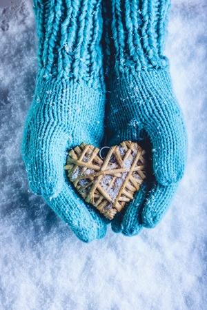 Female hands in light teal knitted mittens with entwined beige flaxen heart on a white snow winter background. Love and St. Valentines Day cozy concept.の写真素材