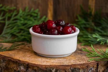 Pickled cranberries in a sweet syrup in a white bowl on the wooden background with the leaves of juniperの写真素材