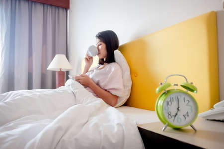 The girl sits in the morning drinking coffee on a bed covered with white linen. There is an alarm...
