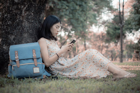 Asian woman tourist A smartphone under the tree. To read social news online. At the public park. It is a relaxing place in vacation lifestyle.の写真素材
