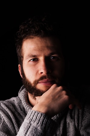 A bearded young man in a sweater stroking his beard looks at the camera on a dark background. The concept of calm and modern men or hipster.の写真素材