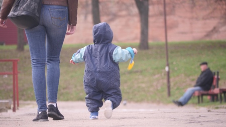 Young woman with her small son who holding baby shovel are walking near a pond in the city park in a spring cloudy day in 4K back view slow motion video.の写真素材