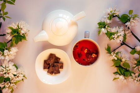 White tea pot, red fruit tea, chocolate, apple tree blossom, white background flat lay. Tea time top view template long web banner. Copy space backdrop mockup design. Floral spring flowers conceptの写真素材