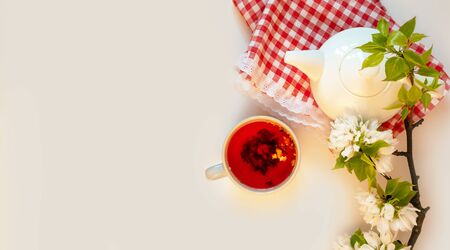 White tea pot, red fruit tea, italian tablecloth, apple blossom, white background flat lay. Tea time top view template long web banner. Copy space backdrop mockup design. Floral spring flowers conceptの写真素材