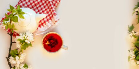 White tea pot, red fruit tea, italian tablecloth, apple blossom, white background flat lay. Tea time top view template long web banner. Copy space backdrop mockup design. Floral spring flowers conceptの写真素材