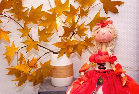 Home concept with interior doll white room, blank wall. Clean wall, light garland, yellow maple leasves. Doll in red dress, sitting on bookshelf. Cozy autumn interior. Space for text, selective focus.の写真素材