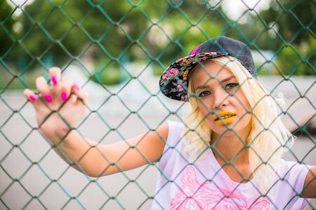 Portrait of a young blond swag girl in cap near the fence.の写真素材