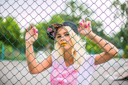 Portrait of a young blond swag girl in cap near the fence.