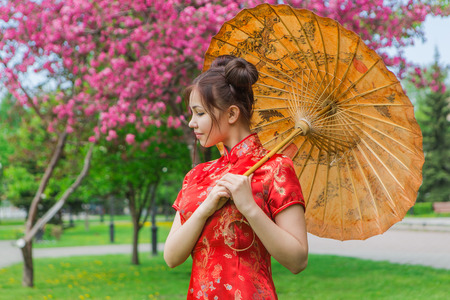 Beautiful asian girl in traditional chinese red dress with bamboo umbrella near pink apple tree in the park.の写真素材