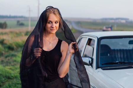 Young girl in long black dress near retro car at sunsetの写真素材