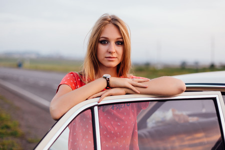 Portrait of a beautiful young woman standing near old car in retro style with bright makeup at sunsetの写真素材