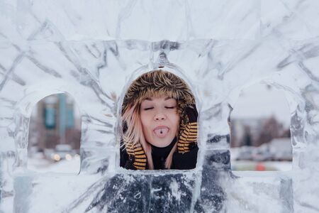 Winter fashion. Portrait of a beautiful young woman in warm clothes outdoor showing tongue near the ice sculpture.の写真素材