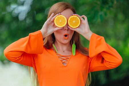 Portrait of pretty red hair woman holding two halves of juicy delicious oranges at summer green park.の写真素材
