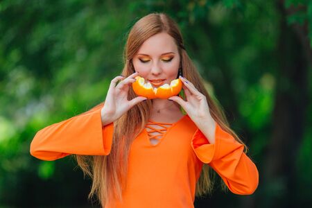 Portrait of pretty red hair woman holding juicy delicious orange at summer green park.の写真素材