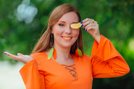 Portrait of pretty red hair woman holding slice juicy delicious orange at summer green park.の写真素材