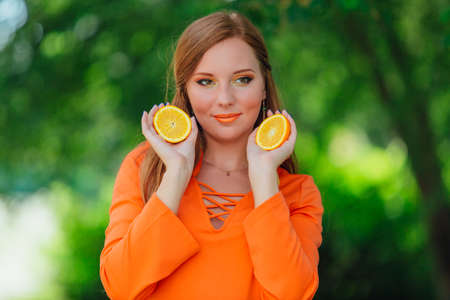Portrait of pretty red hair woman holding two slices of juicy delicious oranges at summer green park.の写真素材