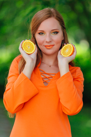 Portrait of pretty red hair woman holding two slices of juicy delicious oranges at summer green park.の写真素材