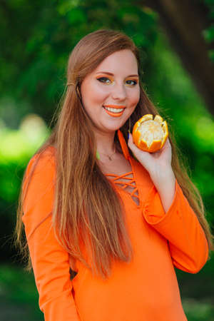 Portrait of pretty red hair woman holding juicy delicious orange at summer green park.の写真素材