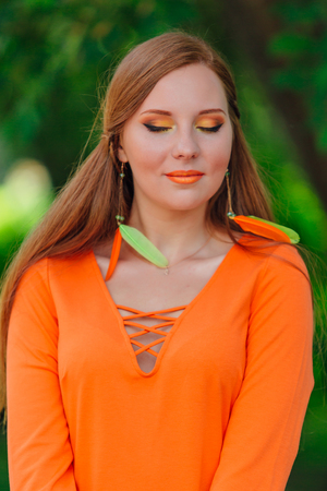 Beautiful romantic red hair girl in orange dress with long feather earrings outdoors.の写真素材