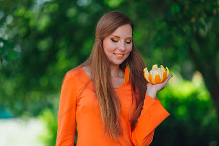 Portrait of pretty red hair woman holding juicy delicious orange at summer green park.の写真素材