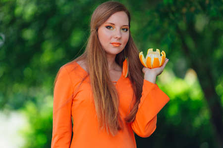 Portrait of pretty red hair woman holding juicy delicious orange at summer green park.の写真素材