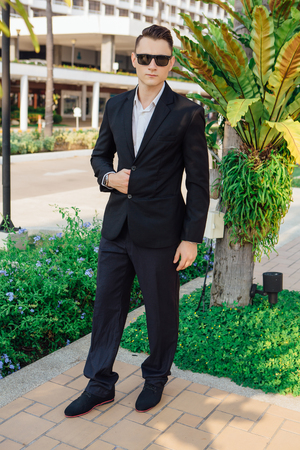Young stylish handsome man posing in modern city park, wearing suit and sunglasses.の写真素材
