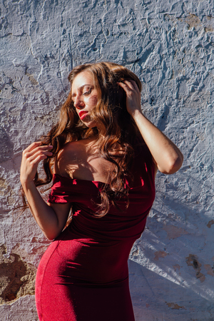 Beautiful young woman in a long red evening gown standing next to the old wallの写真素材