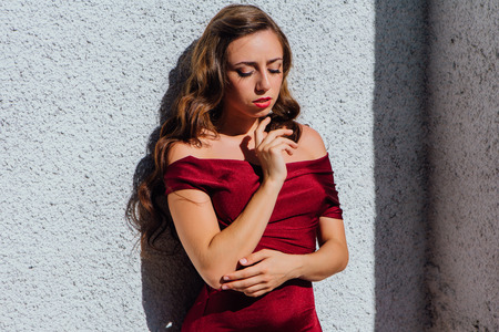 Beautiful young woman in a long red evening gown standing next to the old wallの写真素材