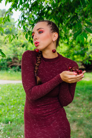 Beautiful young woman with long braid and cherry earrings holding cherry in hands and one cherry in mouthの写真素材