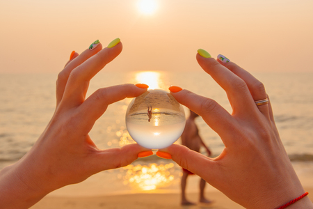 Crystal lens ball in hand with reflection of sea and sunset on the beachの写真素材
