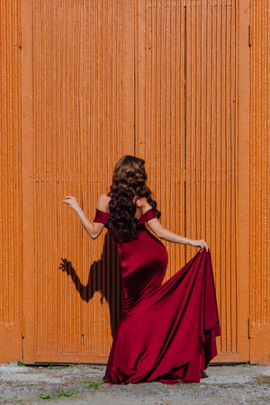 Beautiful young woman in a long red evening gown with a train standing next to the old doorの写真素材