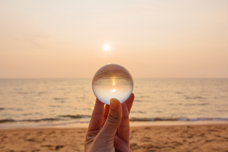Crystal lens ball in hand with reflection of sea and sunset on the beachの写真素材